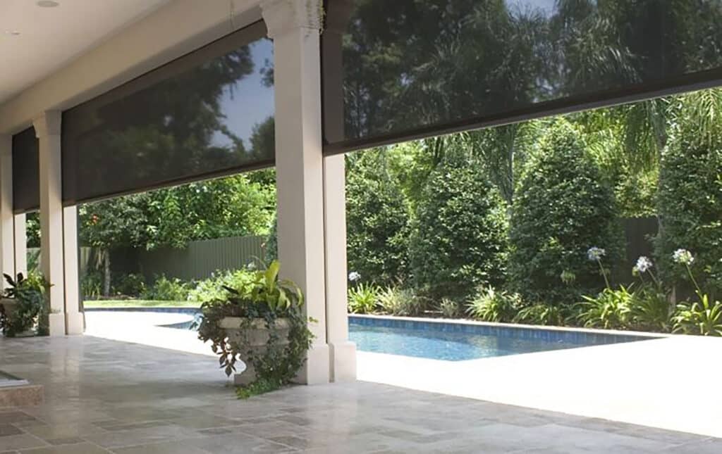 Covered patio with retractable screens partially lowered, overlooking a pool surrounded by lush landscaping